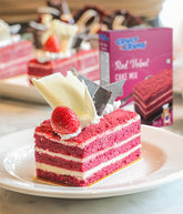 Slice of red velvet cake with cream and strawberry on a plate, with a box of Crust N Crumb's Red Velvet Cake Mix in the background.